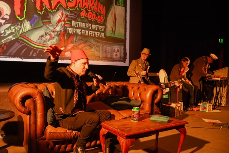 Host speaking on a couch during a live Trasharama A-Go-Go short film festival event on stage with screen showing Australia's nastiest film festival.